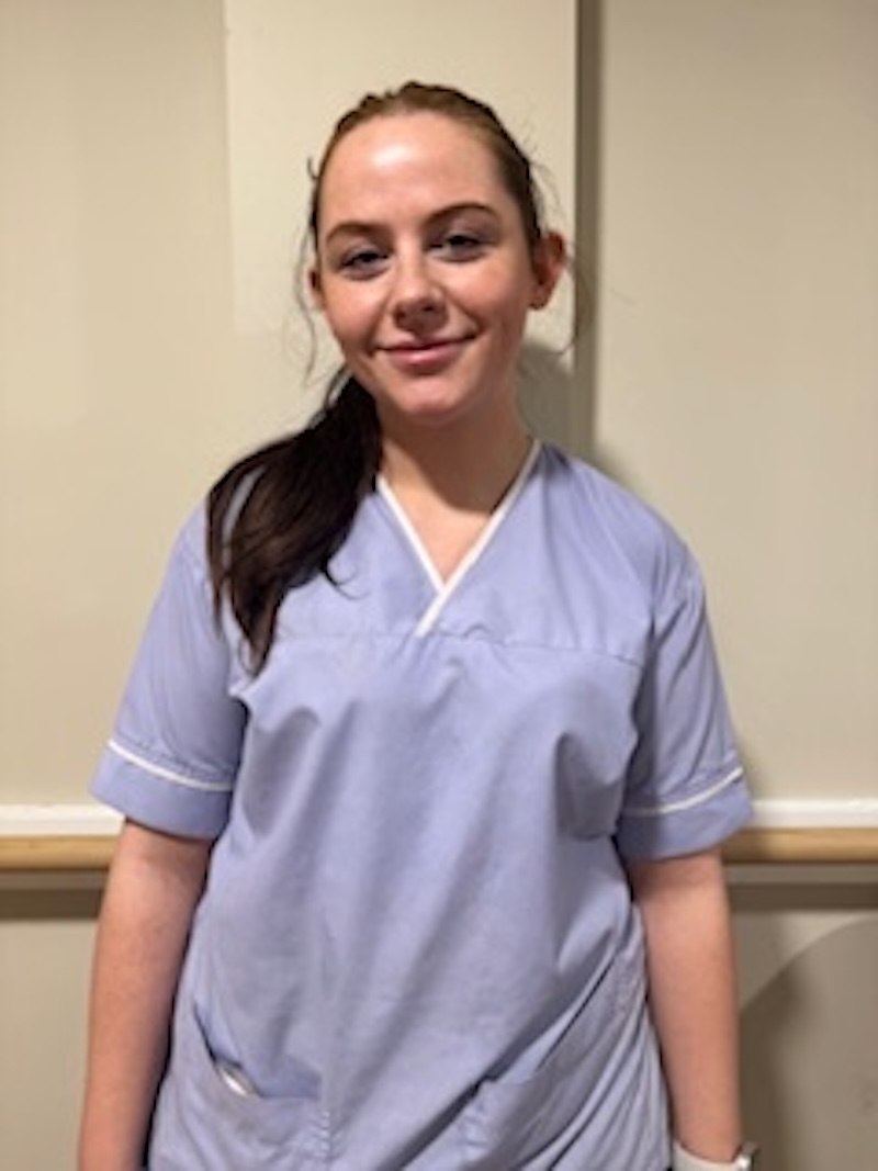 woman with brown hair wearing a lilac uniform stood in front of plain wall having her photo taken for the team page