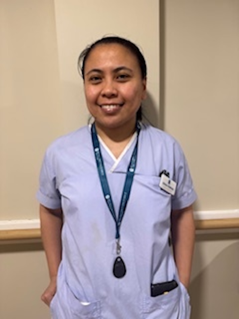 woman with brown hair wearing a lilac uniform stood in front of plain wall having her photo taken for the team page