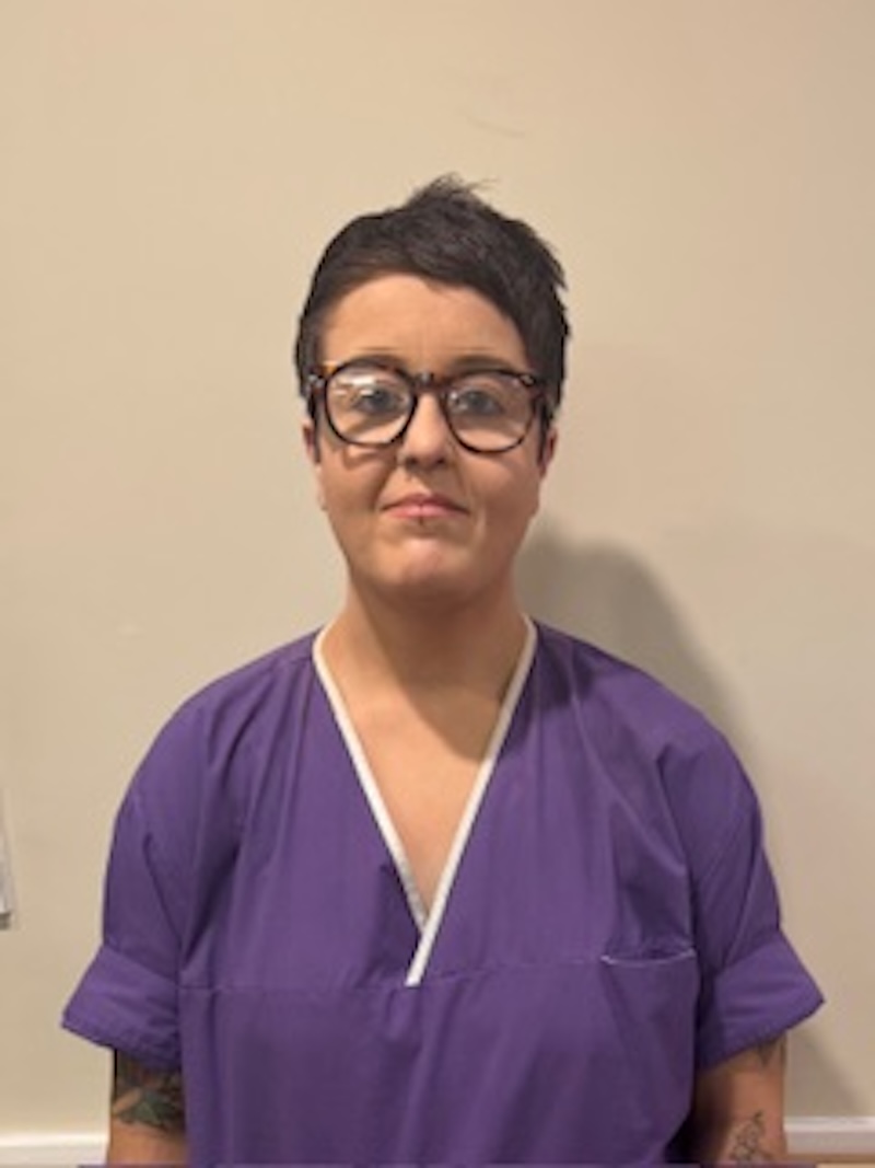 woman with brown hair wearing a purple uniform stood in front of plain wall having her photo taken for the team page