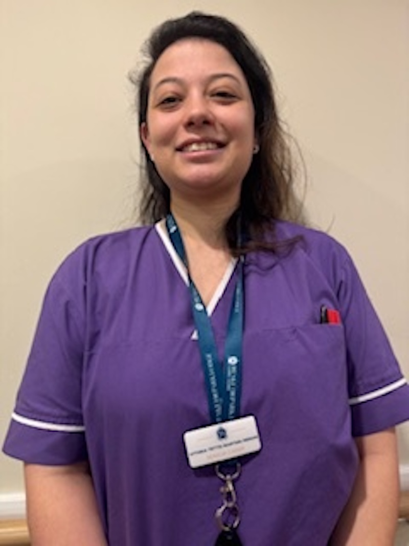 woman with brown hair wearing a purple uniform stood in front of plain wall having her photo taken for the team page